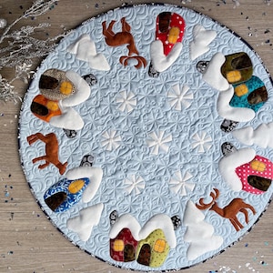 Pode incluir: Uma saia de árvore acolchoada circular com fundo azul claro e costuras de flocos de neve brancos. A borda apresenta renas marrons, árvores brancas cobertas de neve e casas coloridas com padrões e janelas amarelas.
