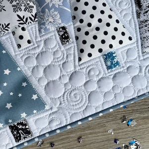 May include: A quilted fabric piece featuring triangular tree shapes in various patterns including black and white snowflakes, blue floral, black polka dots, and dark forest prints, set against a white background with circular and swirl stitching.