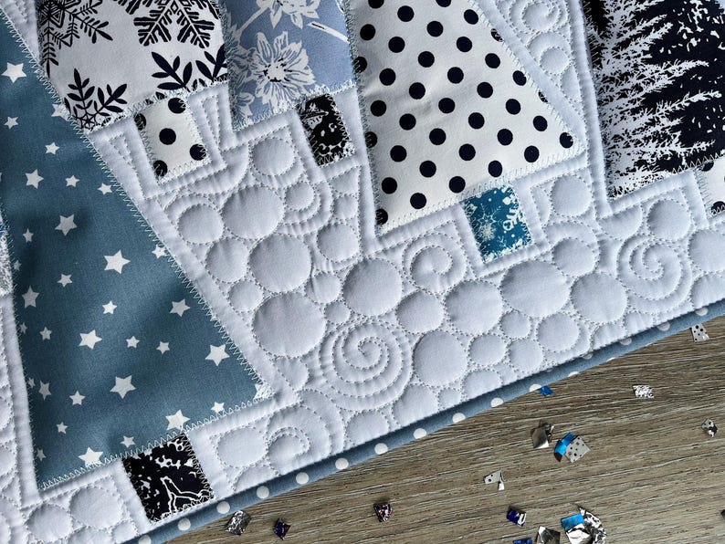May include: A quilted fabric piece featuring triangular tree shapes in various patterns including black and white snowflakes, blue floral, black polka dots, and dark forest prints, set against a white background with circular and swirl stitching.