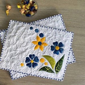 Könnte beinhalten: Zwei weiße, gesteppte Tischsets mit floralem Design. Die Tischsets zeigen gelbe und blaue Blumen mit grünen Blättern. Die Ränder sind mit einem blau-weißen Polka-Dot-Rand versehen. Im Hintergrund befindet sich eine Schüssel mit Pralinen.