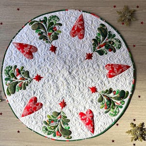 May include: Circular Christmas tree skirt with a white background and a green border. The skirt features red heart and star accents, along with green and red decorative tree-like designs. Gold snowflake and red star embellishments are scattered around the skirt.