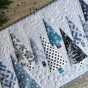 May include: A quilted table runner featuring a series of triangular tree shapes in various shades of blue, white, and black. The fabric patterns include snowflakes, polka dots, floral prints, and geometric designs, all set against a white background.