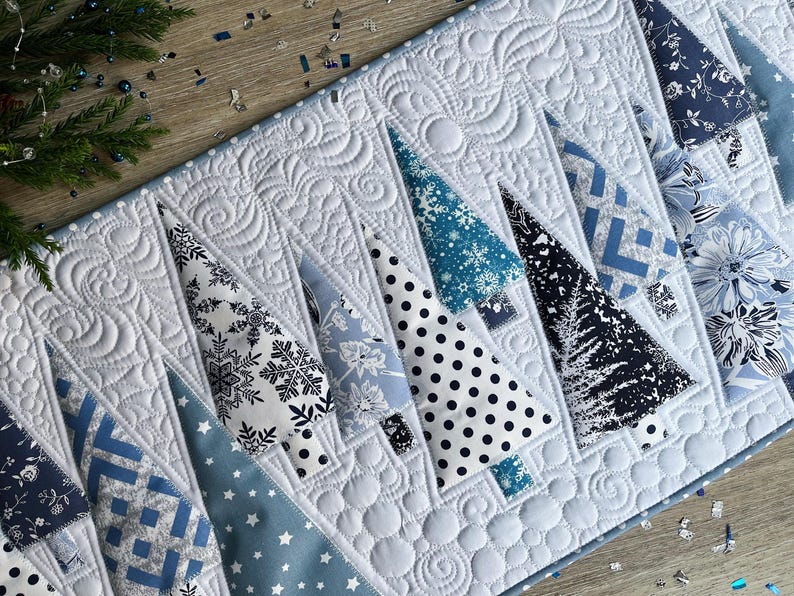 May include: A quilted table runner featuring a series of triangular tree shapes in various shades of blue, white, and black. The fabric patterns include snowflakes, polka dots, floral prints, and geometric designs, all set against a white background.