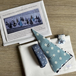 May include: A fabric kit for a quilted table runner titled Winter forest, including white fabric, a blue floral patterned fabric triangle, a blue fabric roll, and a small fabric square with a star print, accompanied by an instructional sheet.