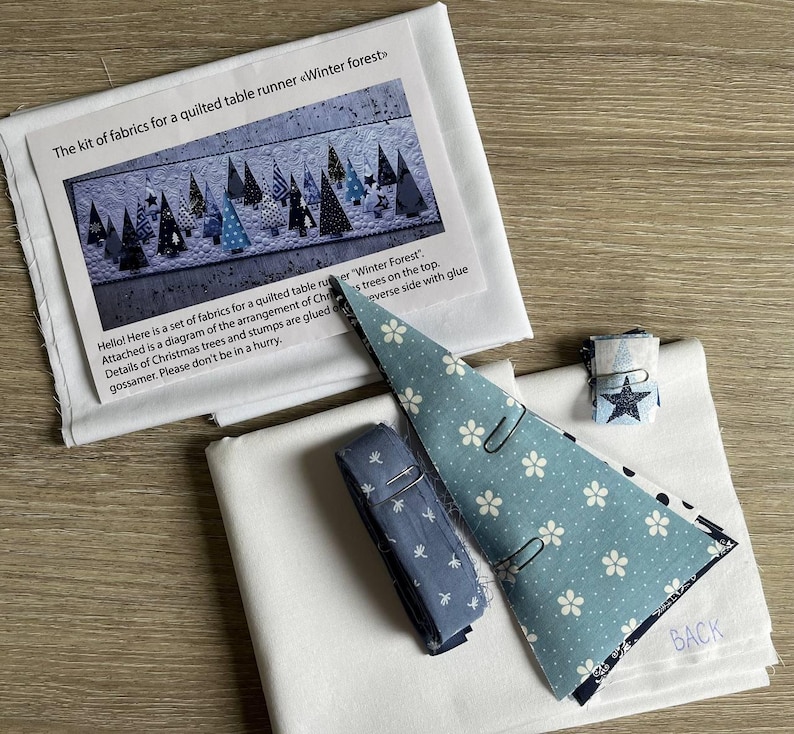 May include: A fabric kit for a quilted table runner titled Winter forest, including white fabric, a blue floral patterned fabric triangle, a blue fabric roll, and a small fabric square with a star print, accompanied by an instructional sheet.