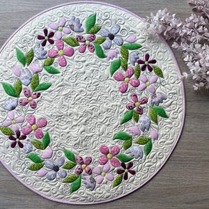 Könnte beinhalten: Ein runder, cremefarbener, gesteppter Tischset mit floralem Design. Das Tischset zeigt einen Kranz aus rosa, lila und dunkelvioletten Blumen mit grünen Blättern. Der Rand ist mit einem hellrosa Rand versehen. Der Hintergrund hat ein detailliertes Steppmuster.