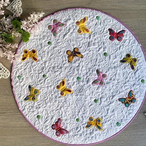 Könnte beinhalten: Ein runder, weiß gesteppter Teppich mit rosa Rand, verziert mit bunten Schmetterlingsapplikationen. Die Schmetterlinge sind in Gelb-, Rosa-, Rot- und Blautönen gehalten und über die Oberfläche verteilt. Grüne Perlen sind ebenfalls vorhanden.