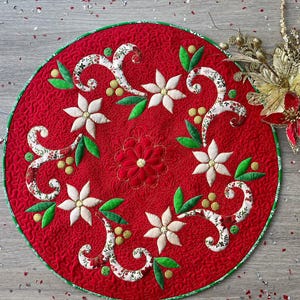 Könnte beinhalten: Eine runde, rote, gesteppte Stoffmatte mit einer gestickten zentralen Blume, umgeben von weißen Blumenapplikationen, grünen Blättern und kleinen gelben Beeren. Die Matte hat einen grünen Rand und ist mit komplizierten Wirbelmustern bestickt.