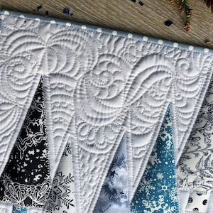 May include: A quilted table runner featuring a white background with intricate swirling stitch patterns. The design includes triangular tree shapes in various shades of black, white, and blue, decorated with snowflake and floral patterns.