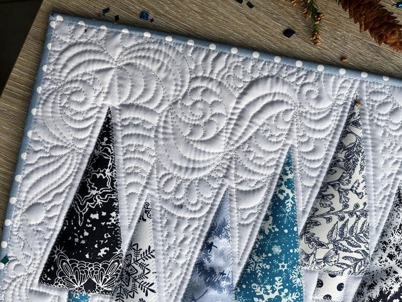 May include: A quilted table runner featuring a white background with intricate swirling stitch patterns. The design includes triangular tree shapes in various shades of black, white, and blue, decorated with snowflake and floral patterns.