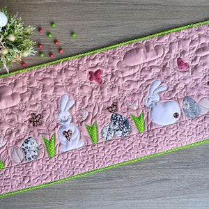 Puede incluir: Camino de mesa de Pascua acolchado rosa con ribete verde. Presenta conejitos blancos aplicados, huevos, mariposas, flores y una pajarera. El camino de mesa está decorado con un estampado floral repetido.