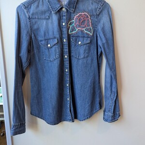 May include: A blue denim shirt with long sleeves, two chest pockets, and a button-down front. The shirt features a colorful embroidered rose patch on the front. The collar has the text "1969".