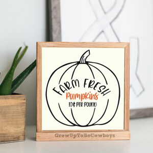 Può includere: Un cartello bianco con un contorno nero di una zucca e il testo "Farm Fresh Pumpkins 10€ al chilo" in lettere arancioni e nere. Il cartello è in una cornice di legno e ha il testo "GrowUpToBeCowboys" in basso.