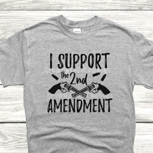 May include: A gray t-shirt with the text "I SUPPORT the 2nd AMENDMENT" printed in black. Two crossed pistols with bullets are depicted above the text.