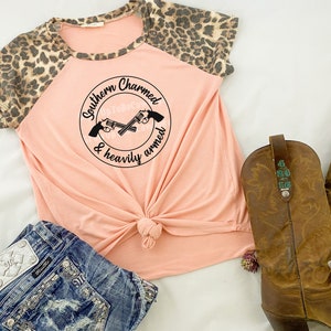 Puede incluir: Una camiseta de color melocot&oacute;n con estampado de leopardo en las mangas. La camiseta tiene un gr&aacute;fico redondo con el texto "Southern Charmed & heavily armed" y un par de pistolas cruzadas.