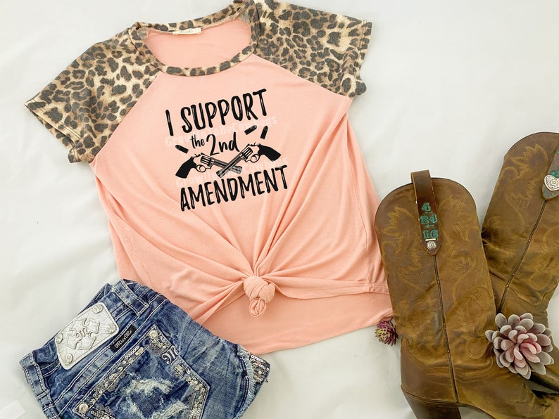 May include: A peach-colored t-shirt with a leopard print on the sleeves. The shirt has a black graphic that says "I Support the 2nd Amendment" with two crossed pistols.
