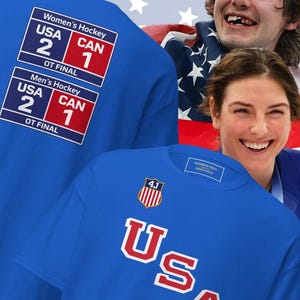May include: Blue t-shirts with "USA" in red and white lettering. The shirts feature the scores of the USA vs. Canada hockey finals, with the USA winning. The text "4JOHNSAPPAREL.COM" is at the bottom.