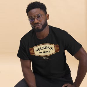 May include: Black t-shirt featuring a rectangular design with the text "NELSON'S RESERVE" in a serif font, with "ROBOSTO EST 1962" below. The design has a brown and gold color scheme. The t-shirt is worn by a person.