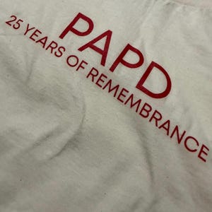 May include: A white fabric garment featuring red text printed diagonally across the surface. The text reads PAPD 25 YEARS OF REMEMBRANCE in a bold, sans-serif font. The material appears to be a soft, woven textile with a visible weave pattern.