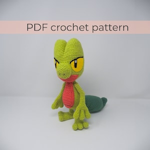 Puede incluir: Patrón de crochet para un personaje Pokemon verde y rojo. El personaje tiene ojos amarillos y una cola larga. El patrón está disponible para descargar en PDF.