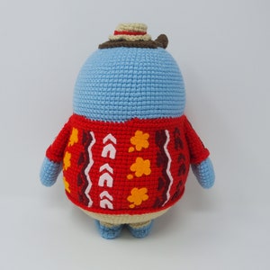 May include: A blue crocheted stuffed animal wearing a red and brown sweater with a white and yellow pattern. The animal is wearing a brown and beige hat.