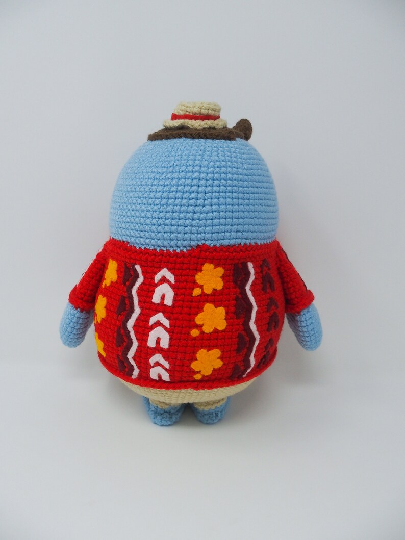 May include: A blue crocheted stuffed animal wearing a red and brown sweater with a white and yellow pattern. The animal is wearing a brown and beige hat.