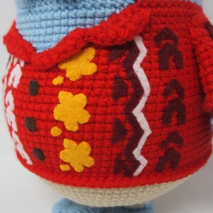 May include: A crocheted stuffed animal wearing a red sweater with yellow and brown details. The sweater has a white zig-zag pattern.