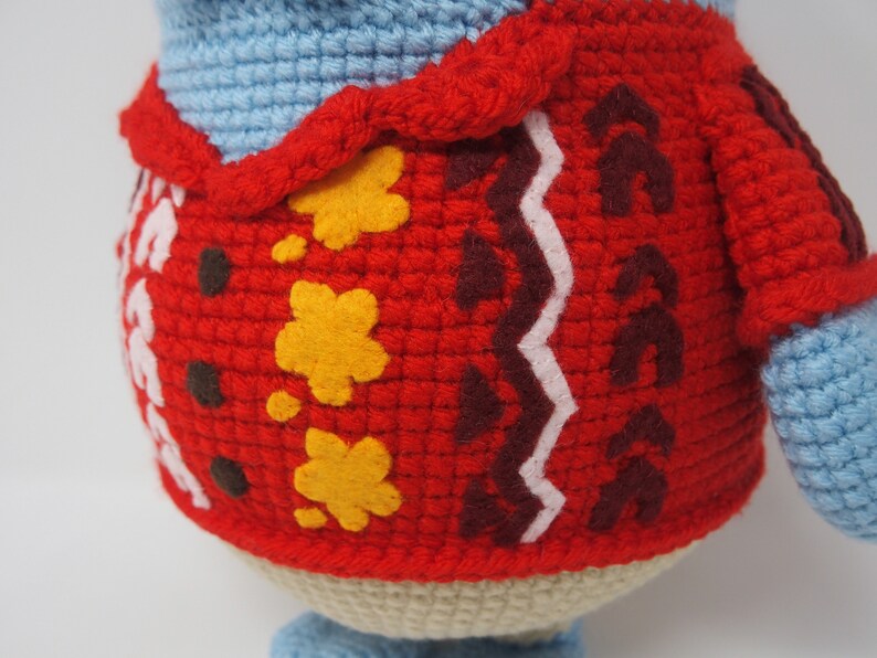 May include: A crocheted stuffed animal wearing a red sweater with yellow and brown details. The sweater has a white zig-zag pattern.