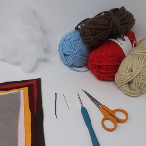 May include: A collection of craft supplies for a DIY project, including yarn in brown, blue, red, and beige, a needle, a crochet hook, a pair of scissors, felt fabric in various colors, and stuffing.