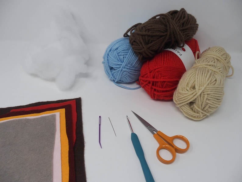 May include: A collection of craft supplies for a DIY project, including yarn in brown, blue, red, and beige, a needle, a crochet hook, a pair of scissors, felt fabric in various colors, and stuffing.