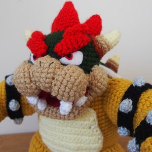 May include: A handmade Bowser plushie, crocheted with yellow, tan, red, green, and black yarn. The plush features Bowser's iconic spiky shell, horns, and facial features, including a wide open mouth with white teeth. The toy is a representation of the video game character.