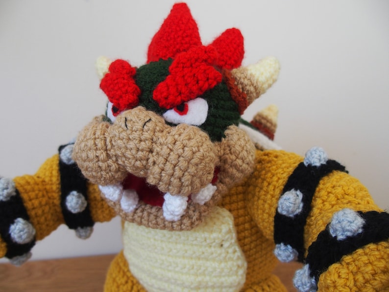 May include: A handmade Bowser plushie, crocheted with yellow, tan, red, green, and black yarn. The plush features Bowser's iconic spiky shell, horns, and facial features, including a wide open mouth with white teeth. The toy is a representation of the video game character.