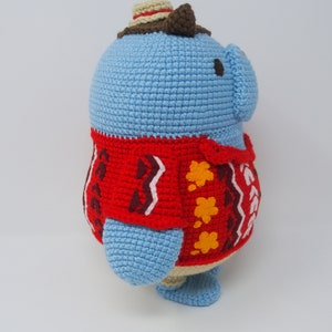 May include: A blue crocheted stuffed animal wearing a red and brown sweater with a brown hat. The animal has a round body and small legs.