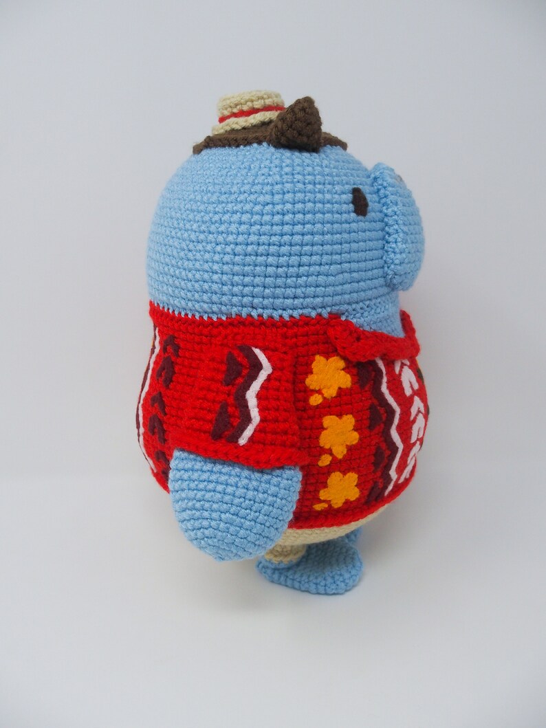 May include: A blue crocheted stuffed animal wearing a red and brown sweater with a brown hat. The animal has a round body and small legs.