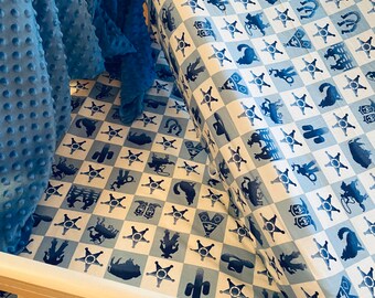 May include: A white crib with a blue and white checkered crib sheet featuring a repeating pattern of cowboy boots, stars, horseshoes, and cacti. The crib sheet is tucked into the crib.