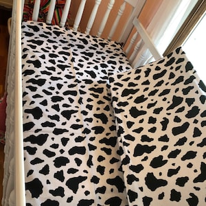 May include: A white crib with a black and white cow print fitted sheet and a black and white cow print crib bumper.