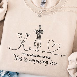 May include: A beige sweatshirt with a black line drawing of a cross, nails, and a heart. The text "THIS IS AMAZING GRACE. This is unfailing love." is below the drawing. A silver necklace is draped across the sweatshirt.