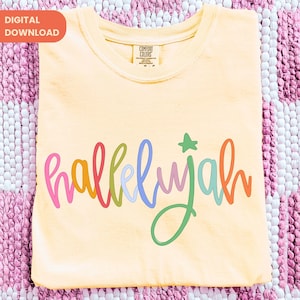 May include: A light yellow t-shirt with the word "hallelujah" in colorful script across the front. The word is in pink, orange, green, blue, and purple. A green star is above the letter "j". The shirt is folded on a pink and white patterned surface.