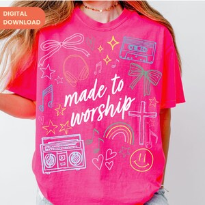 Op de afbeelding: Felroze t-shirt met de tekst "made to worship" in witte letters. Het shirt heeft verschillende doodle-achtige afbeeldingen, waaronder sterren, muzieknoten, een cassettebandje, een koptelefoon en een regenboog.