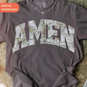 Op de afbeelding: Donkergrijs T-shirt met het woord "AMEN" in een camouflage-lettertype. Het shirt heeft een ronde hals en lange mouwen. Een delicate zilveren ketting met een vlinderhanger is zichtbaar bij de halslijn. De tekst "DIGITALE DOWNLOAD" staat in een oranje kader linksboven.
