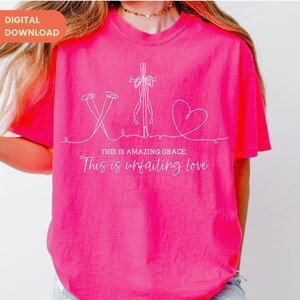 以下が含まれることがあります： 明るいピンク色のTシャツ。白い線で描かれた釘、十字架、ハートのイラストが特徴です。「THIS IS AMAZING GRACE. This is unfailing love」というテキストがイラストの下にあります。Tシャツは半袖です。