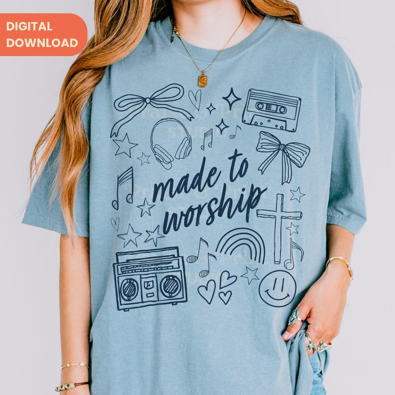 Op de afbeelding: Lichtblauw T-shirt met de handgeschreven tekst "made to worship". Het shirt heeft doodle-achtige illustraties van muzieknoten, sterren, een cassettebandje, een boombox, een kruis, een regenboog en een smiley.