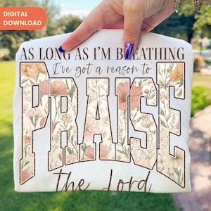 May include: White t-shirt with the text "AS LONG AS I'M BREATHING I've got a reason to PRAISE The Lord." The word "PRAISE" is in large, block letters filled with a floral pattern in shades of peach, cream, and brown. The words are in a brown, vintage font.
