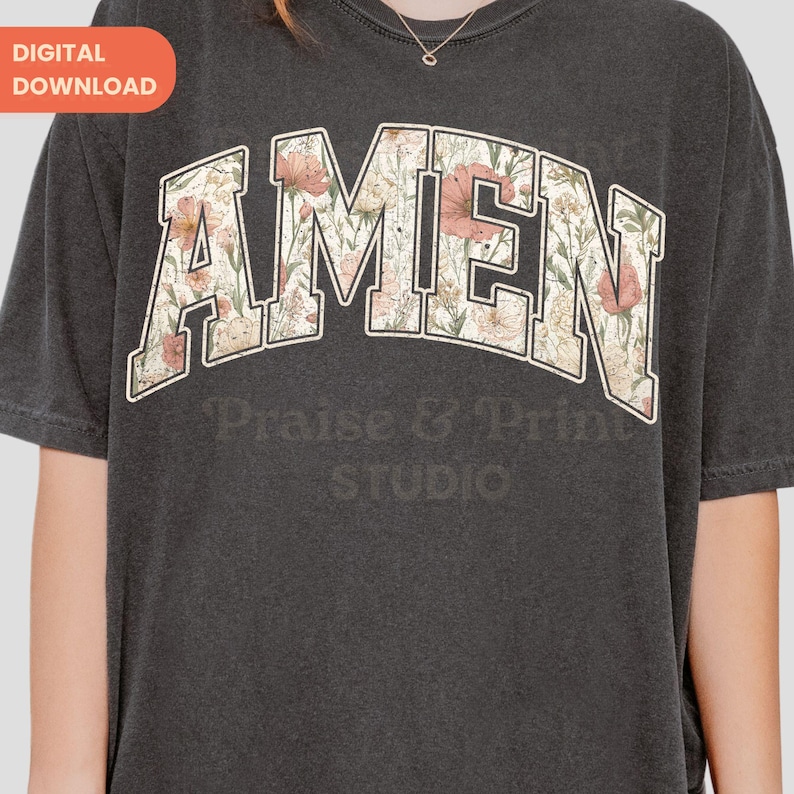May include: Dark gray t-shirt featuring the word "AMEN" in a floral design. The text is in a collegiate-style font with a white outline. Below the word "AMEN" is the text "Praise & Print STUDIO".