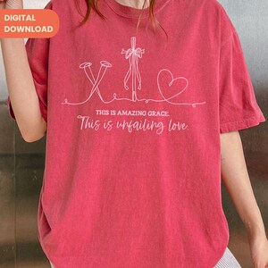 以下が含まれることがあります： 釘、十字架、ハートが描かれた白いグラフィックデザインの赤いTシャツ。「THIS IS AMAZING GRACE. This is unfailing love」の文字が下にプリントされています。シャツは半袖です。