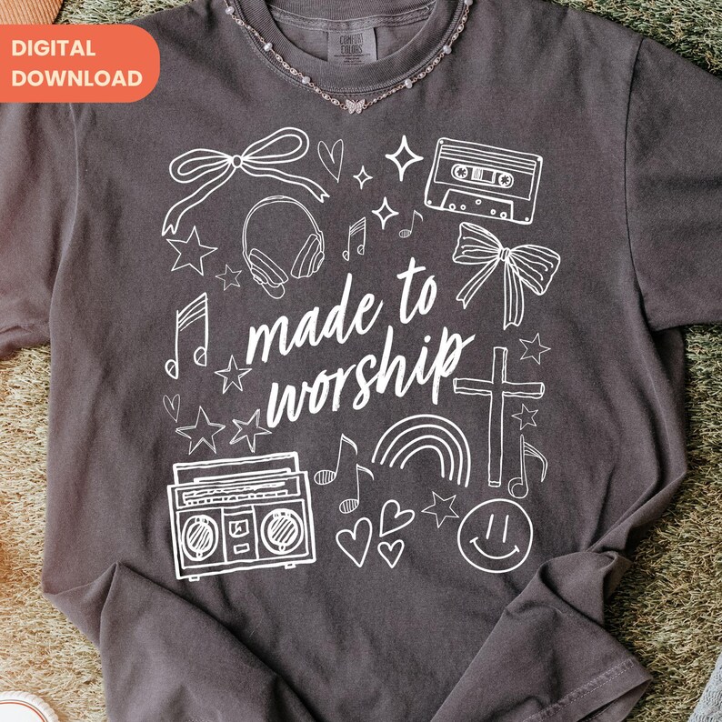 Op de afbeelding: Donkergrijs t-shirt met een wit grafisch ontwerp. Het ontwerp bevat een strik, koptelefoon, muzieknoten, sterren, een cassettebandje, een boombox, een kruis, een regenboog, harten en de tekst "made to worship".