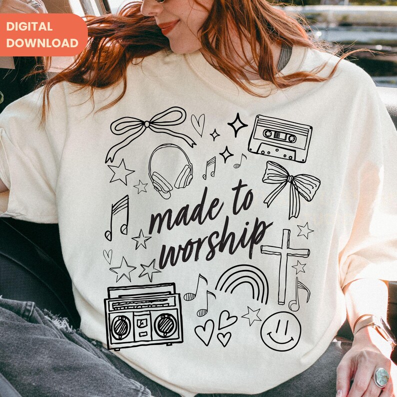 Op de afbeelding: Cr&egrave;mekleurige sweatshirt met een zwart grafisch ontwerp. Het ontwerp bevat een strik, koptelefoon, muzieknoten, sterren, een cassettebandje, een kruis, een regenboog, een boombox, harten en de tekst "made to worship".