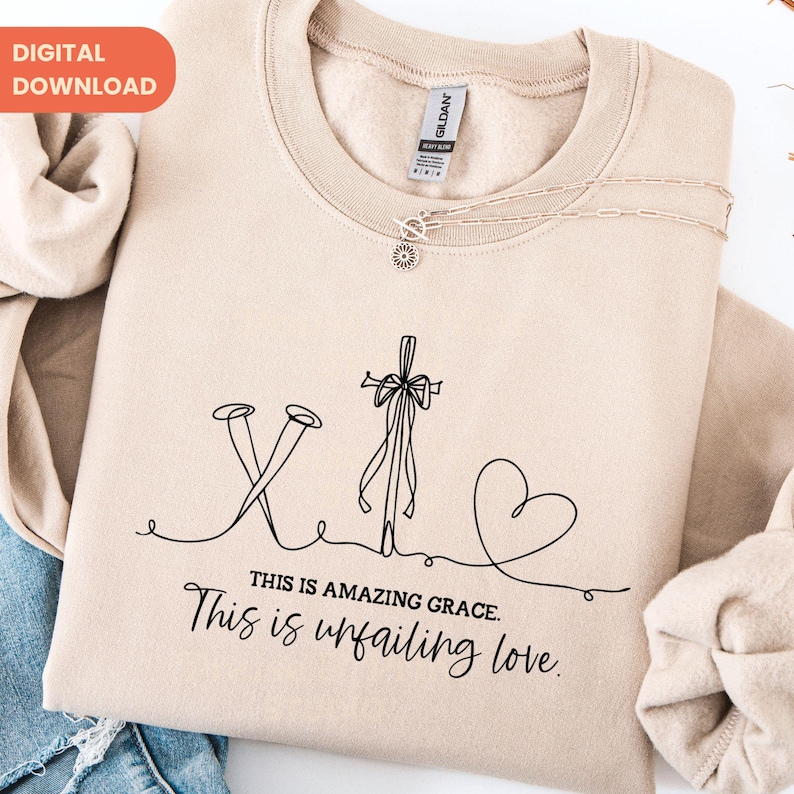 May include: A beige sweatshirt with a black line drawing of a cross, nails, and a heart. The text "THIS IS AMAZING GRACE. This is unfailing love." is below the drawing. A silver necklace is draped across the sweatshirt.