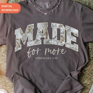 Op de afbeelding: Donkergrijs t-shirt met de tekst "MADE for more" in een collegiale lettertype, met een camouflage-achtig patroon. De zin "Ephesians 2:10" staat eronder. Een delicate ketting is bij de halslijn.