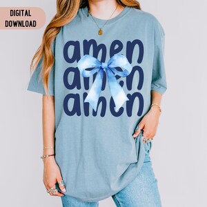 Op de afbeelding: Een lichtblauw T-shirt met het woord "amen" drie keer herhaald in donkerblauwe letters. Een blauwe en witte aquarel strik is gecentreerd op het T-shirt.